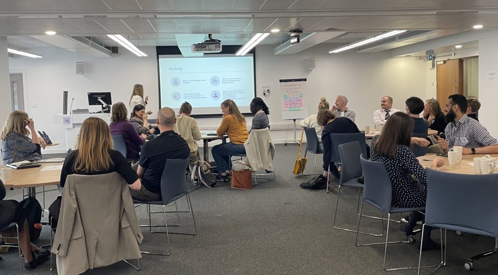 Photo of roundtable discussion. Groups of people sitting around circular tables in front of a Powerpoint presentation being given by Maaya Modha-Patel,