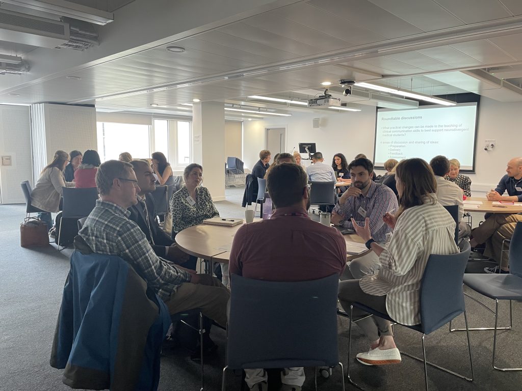 Group of people in conversation sat round a table. Whiteboard in the background showing roundtable topic for discussion.