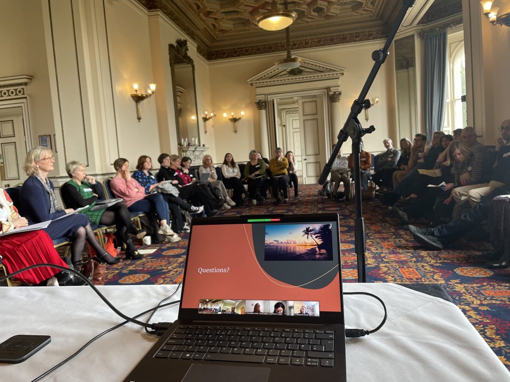 Group of UKCCC members at a meeting, sat in horse shoe shape in front of a laptop with a slide that has the word 'Questions?' written on it.